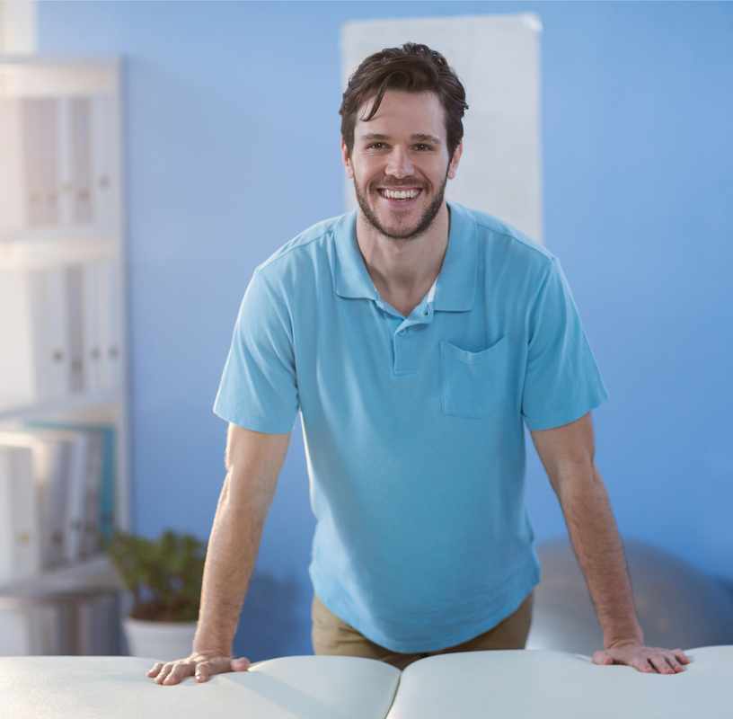 Uśmiechnięty fizjoterapeuta, ubrany w niebieski uniform, stojący w gabinecie rehabilitacyjnym, oparty o stół rehabilitacyjny i patrzący w obiektyw.